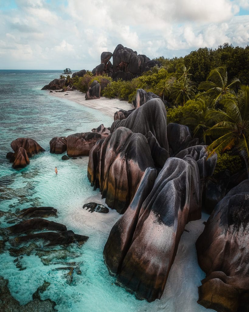 Aerial view of personal standing in clear blue water surrounded by tall rock formations at Anse Source d'Argent beach, La Digue