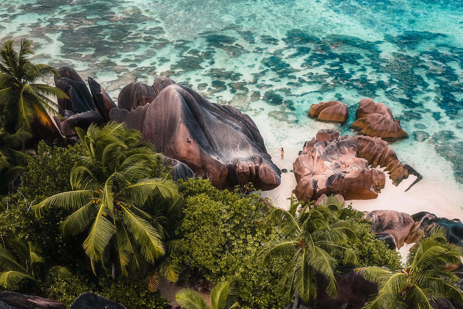 Aerial drone view of a woman standing on a tropical beach with palm tress and rock formations at Anse Source d'Argent, La Digue