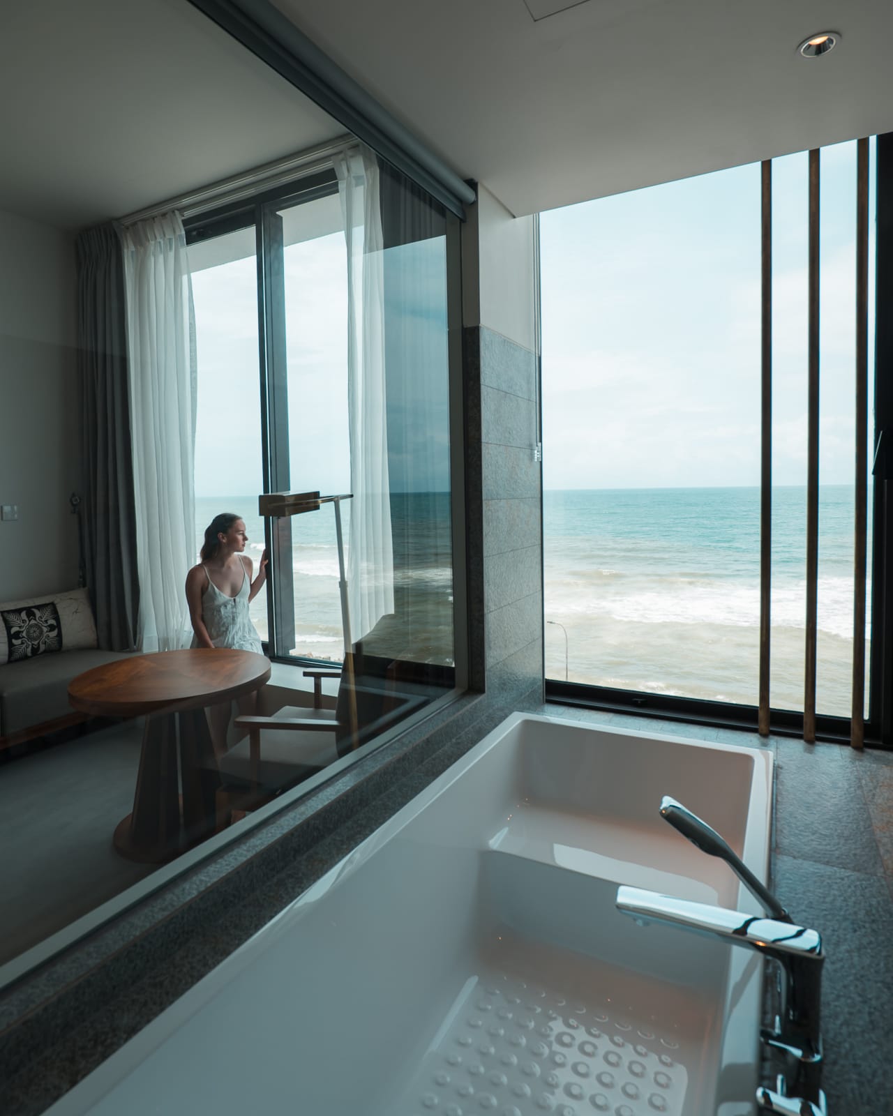 woman sitting on chair in hotel room with bathtub looking out over ocean 