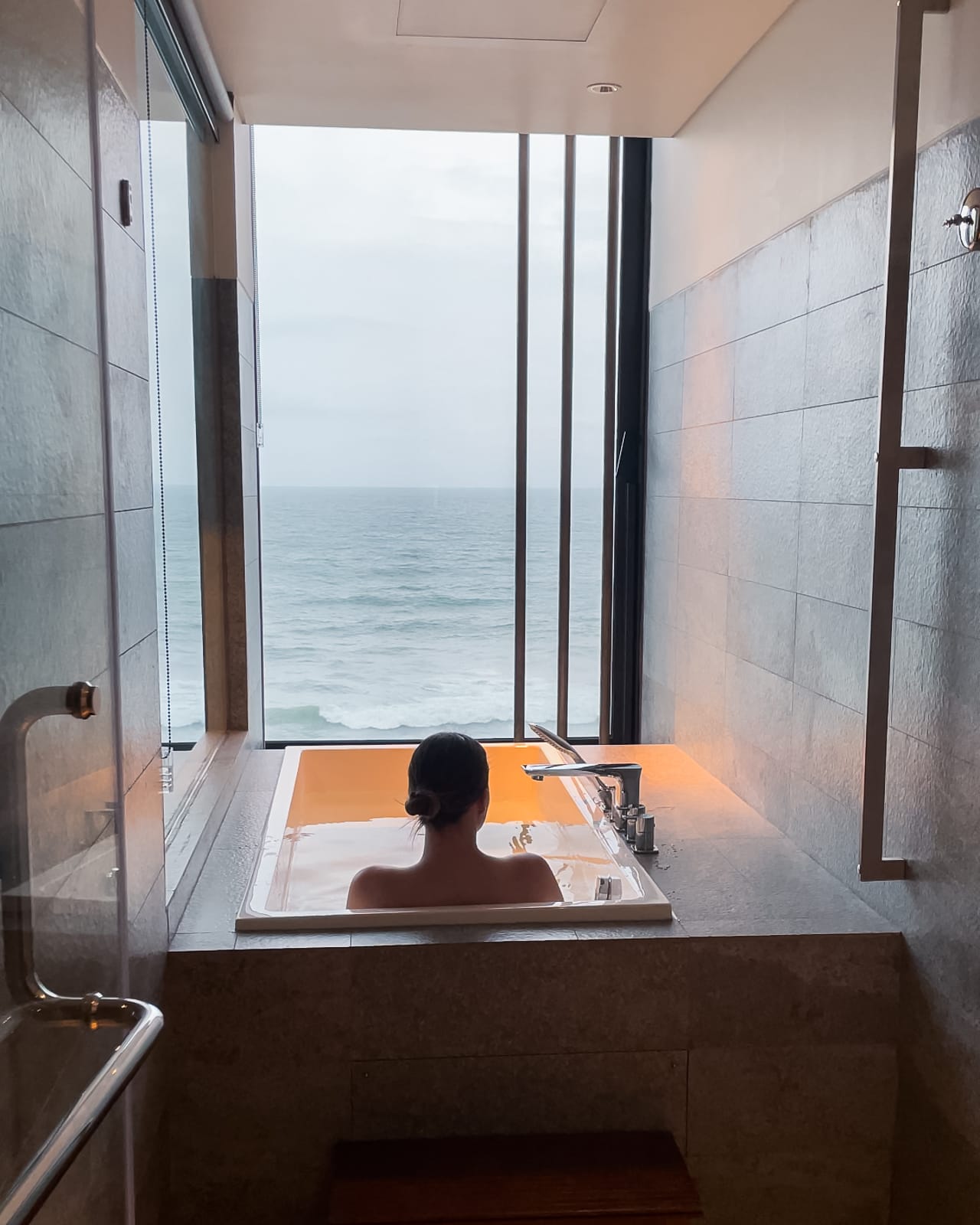 woman in bathtub looking out over ocean through window