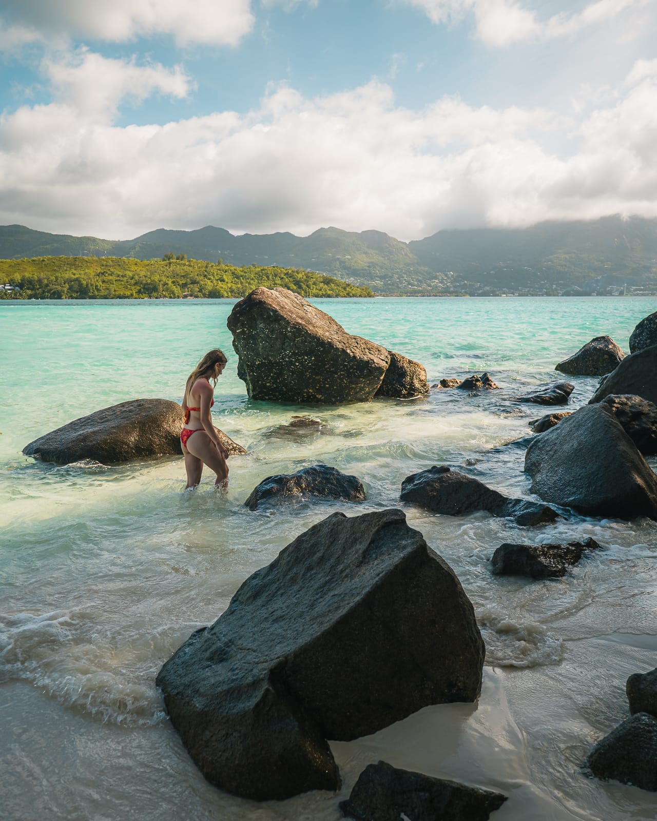 woman standing on rocky beach with turqoise blue water and tropical mountains 