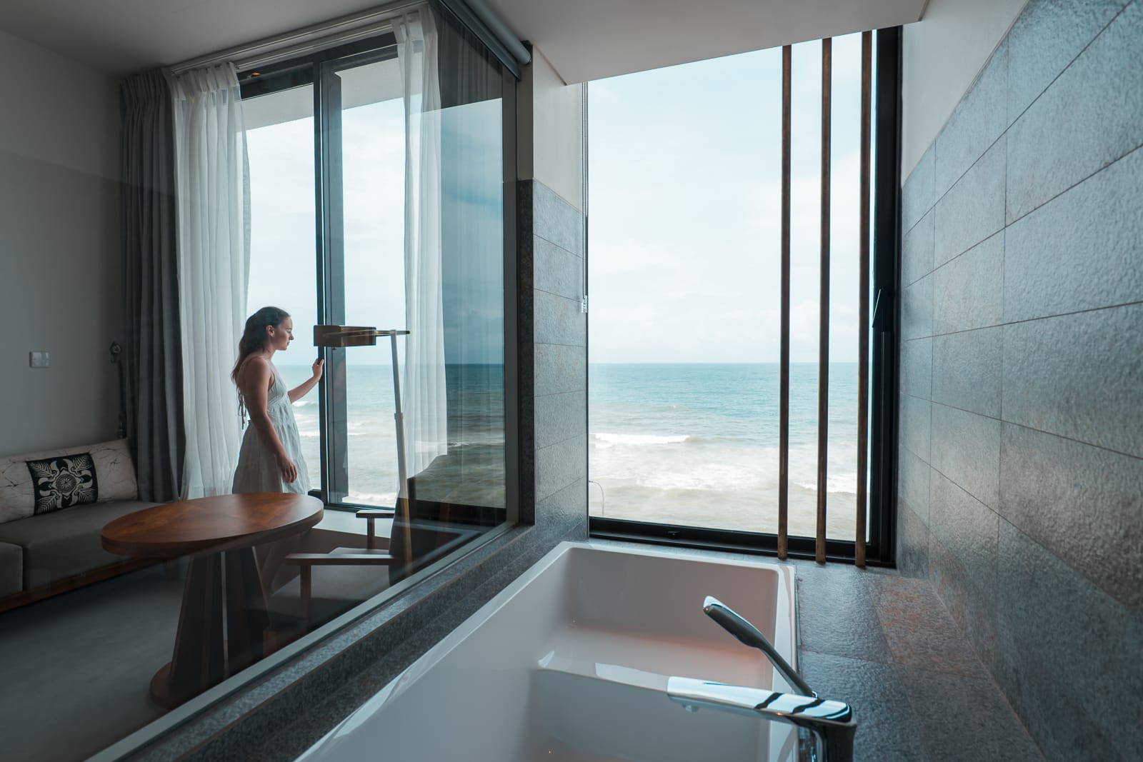 woman standing in hotel room with bathtub and ocean view from window 